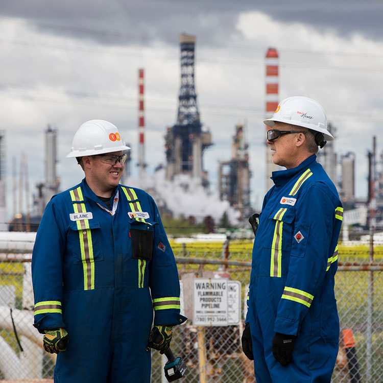 Two pipeline technicians in blue coveralls and wearing personal protective equipment at a facility site talking to each other.