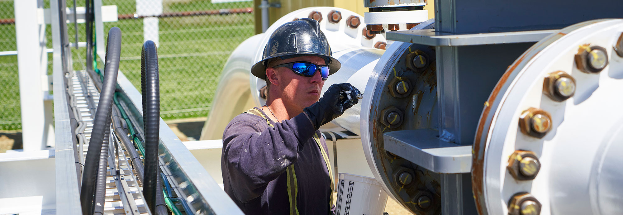 A man wearing a safety hat, sunglasses, work gloves, and high-visibility shirt painting an above-ground pipeline segment with white paint.