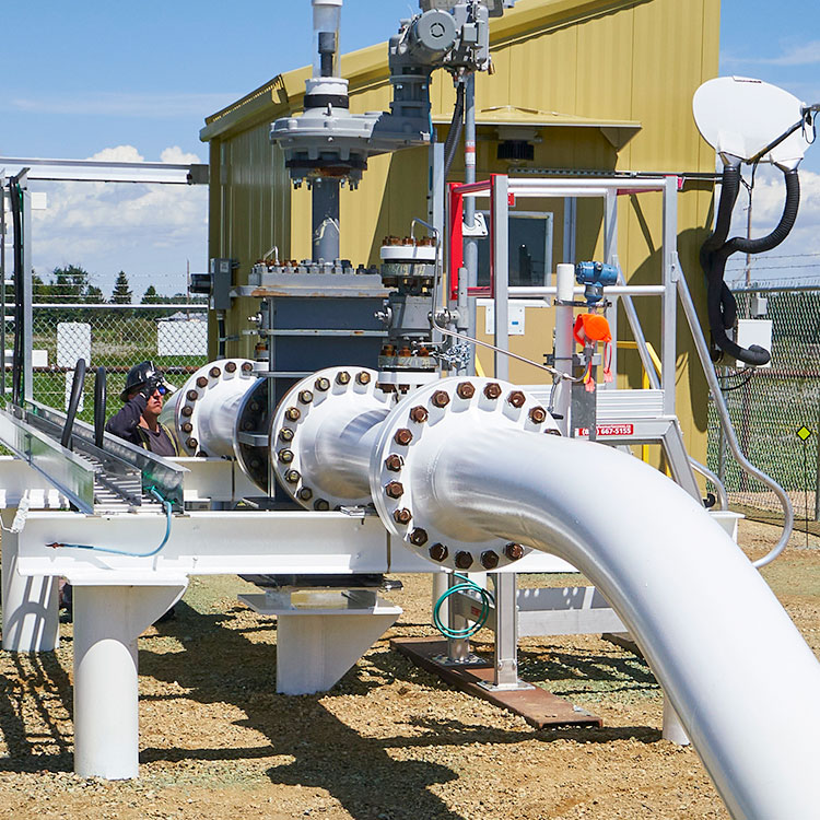 A pipeline segment, painted white, comes above ground at a pumping station, with a yellow building behind the segment within the fenced station.