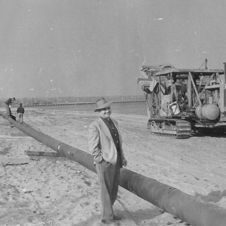 Black-and-white image of a man in a suit coat and hat standing next to a pipeline segment, with and old excavator nearby.