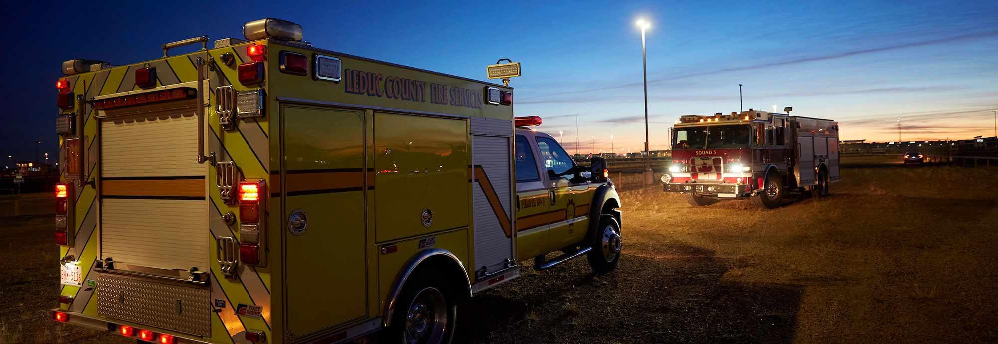 A yellow fire services vehicle faces a red fire truck parked in a field near a highway at dawn.