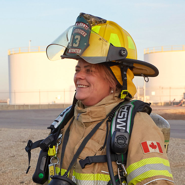 A female firefighter in full firefighting gear smiles, standing near a fuel storage tank farm.