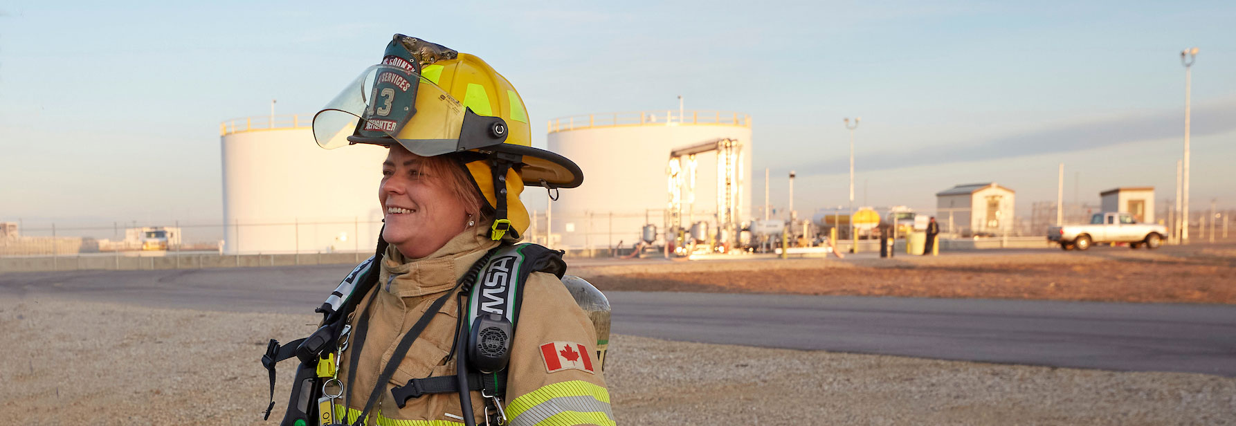 A female firefighter in full firefighting gear smiles, standing near a fuel storage tank farm.