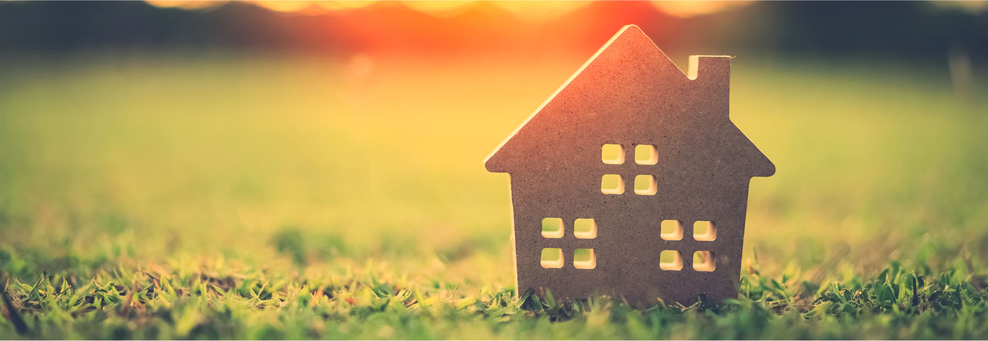 A cutout of a little house is situated on a lawn, with the sun rising in the background.