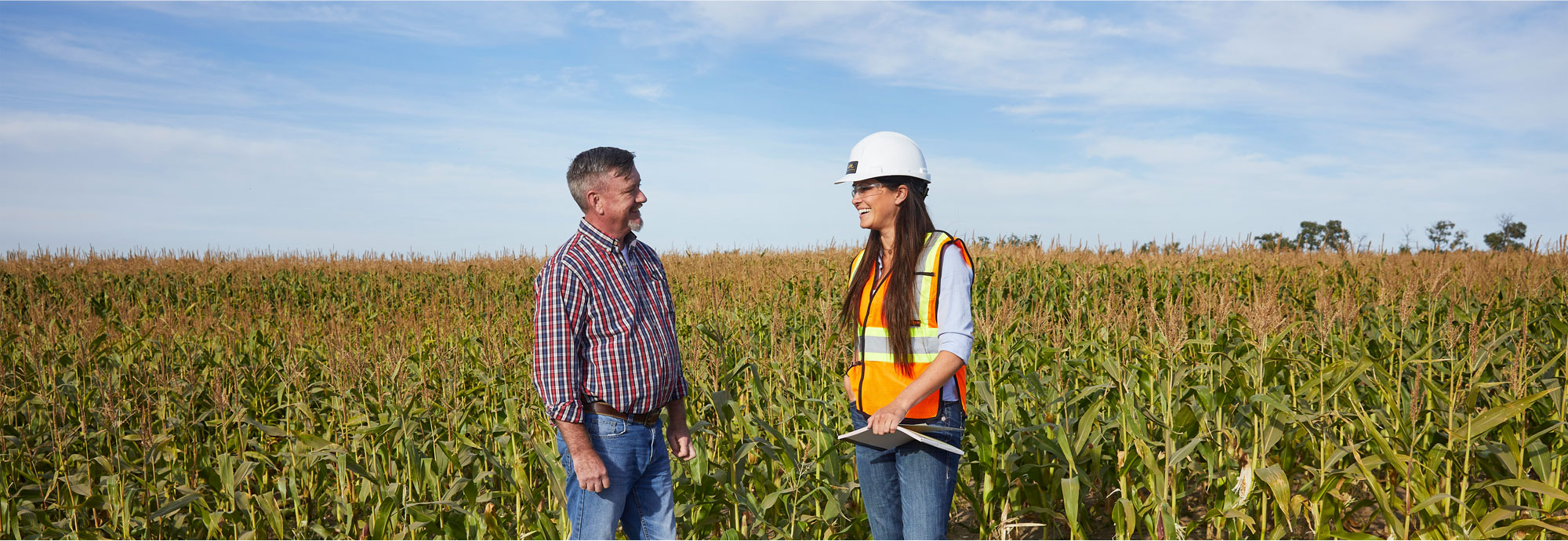 A man stands in a field on the left smiling to a woman wearing a high-visibility vest and hard hat, holding a notebook, and looking back at the man smiling.