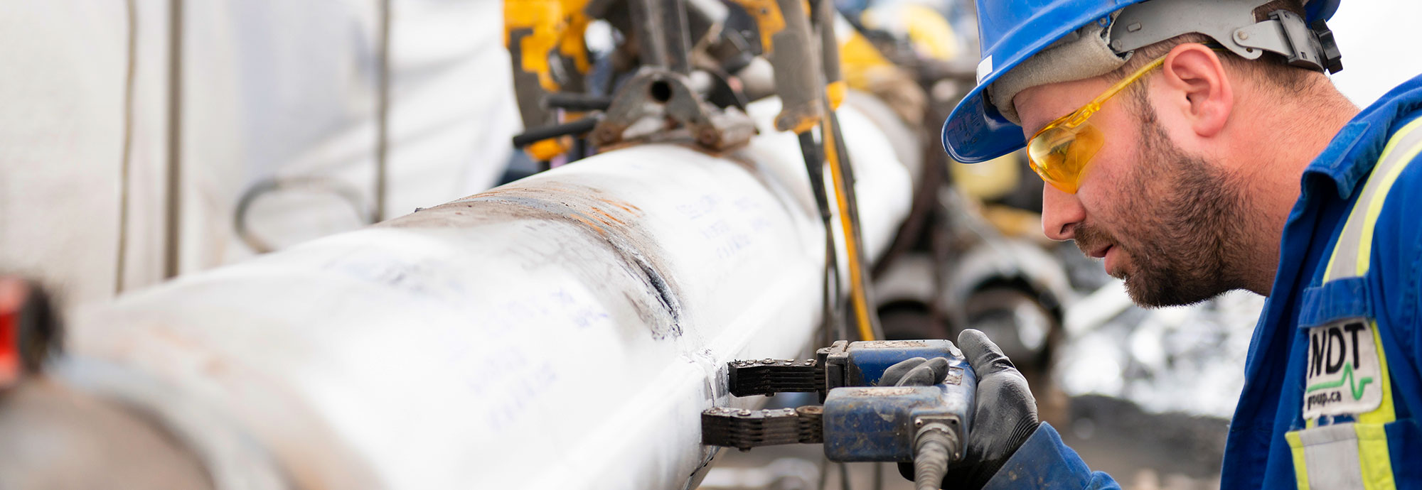 A pipeline technician wearing a safety hat, safety glasses, and work gloves holds a tool against a pipeline sleeve.