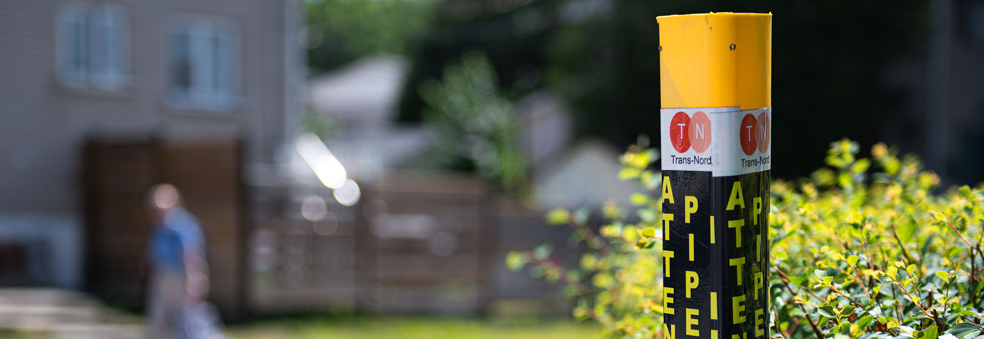 A yellow TNPI pipeline marker near a hedge with a fence and yard in the background in Laval, Quebec.