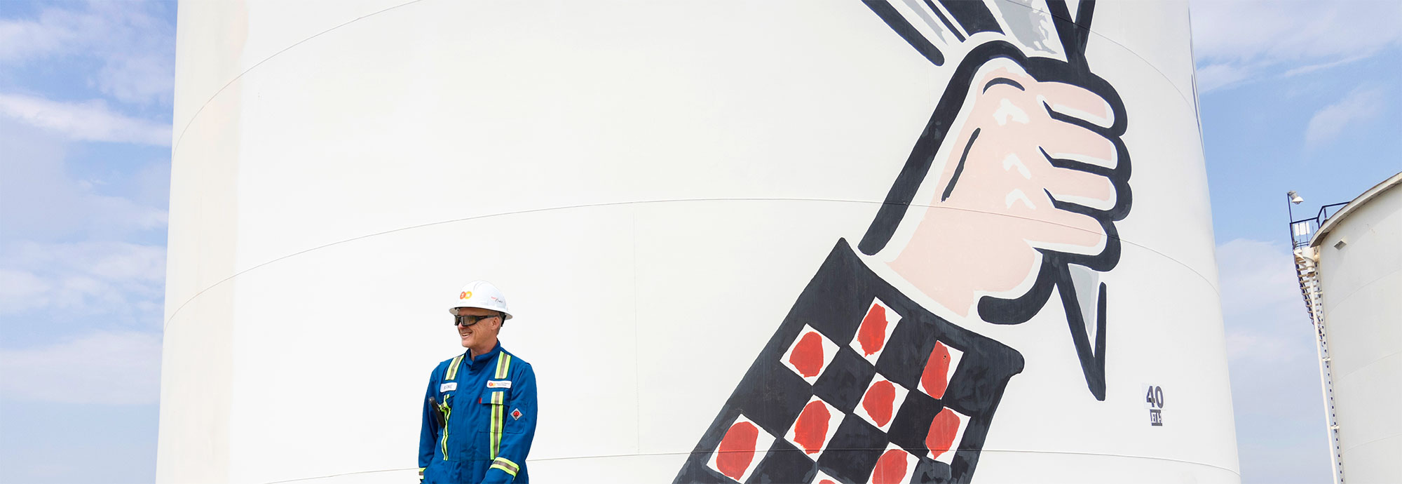 A TNPI employee wearing blue coveralls, a hard hat and safety glasses walks in front of a fuel storage tank in Calgary.