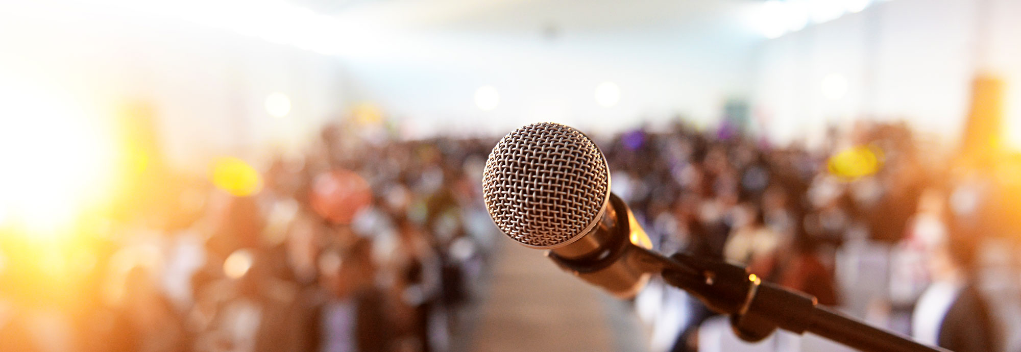 A microphone is in focus, with an out-of-focus crowd of people seated in front.